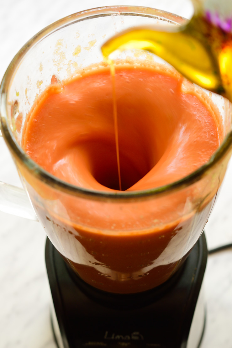 Drizzling olive oil into a blender of gazpacho.