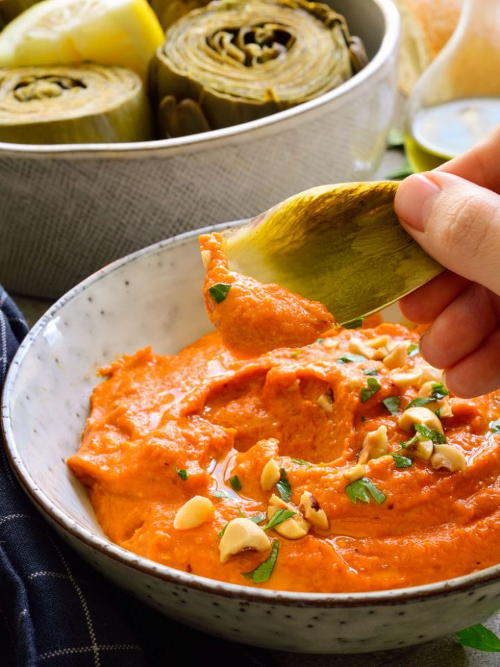 Romesco sauce in a bowl with an artichoke leaf being dipped in.