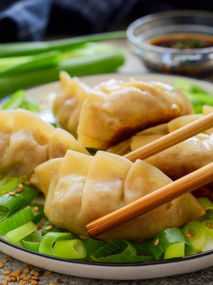 A plate of vegan dumplings. The front one is held by chopsticks. Dipping sauce in the back.